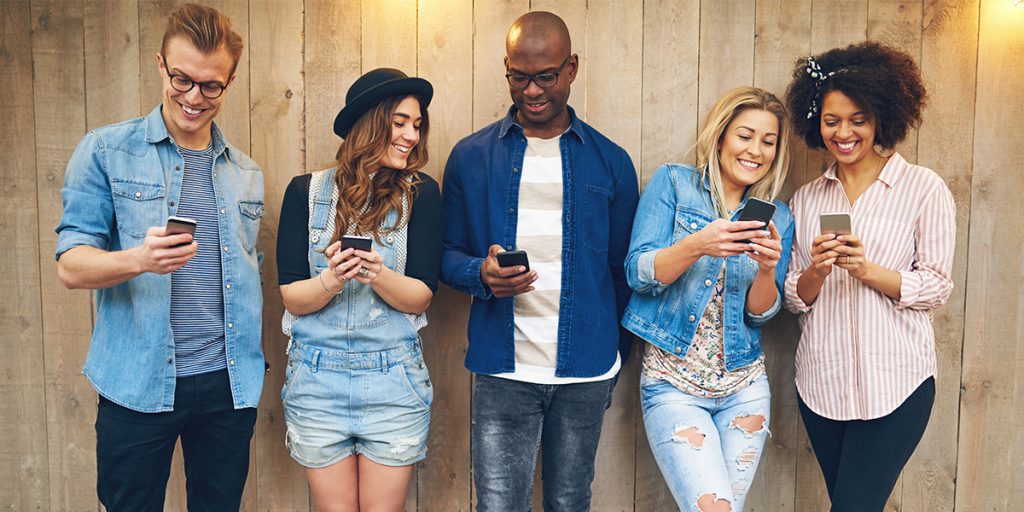 two men and three women stand along a wood fence all looking at their mobile phones